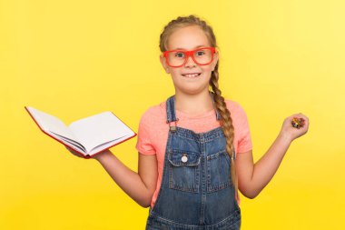 Yaratıcı öğrenme konsepti. Gözlüklü, elinde kalem ve defter olan, kameraya gülümseyen, ilginç eğitimin keyfini çıkaran zeki küçük bir kızın portresi. Stüdyo çekimi sarı arkaplanda izole edildi