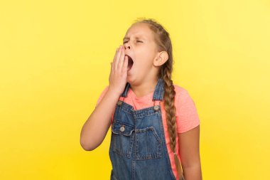 Yorgun bir çocuğun portresi, kot pantolonlu, esnemiş, bitkin, uyku problemleri ve enerji eksikliği hisseden uykulu küçük bir kız. Sarı arka planda izole edilmiş kapalı stüdyo çekimi