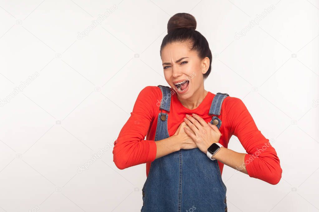 Cardiac problems. Portrait of young fatigued woman with hair bun in denim overalls clasping chest, feeling acute pain, heart attack from anxiety, infarction. studio shot isolated on white background