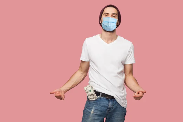 Confident cool young hipster man with surgical medical mask in white shirt and casual hat standing, hold money on the pocket, and pointing finger on it. Indoor, isolated, studio shot, pink background