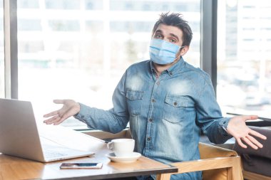 Not sure! Portrait of confused young man with medical mask in blue shirt sitting, working on laptop with raised arms and puzzled face, looking at camera. indoor working and health care concept. clipart
