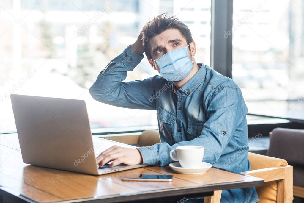 businessman with surgical medical mask in blue shirt sitting and typing on laptop, having new idea and planning own strategy, holding one hand on the head. indoor working and health care concept.