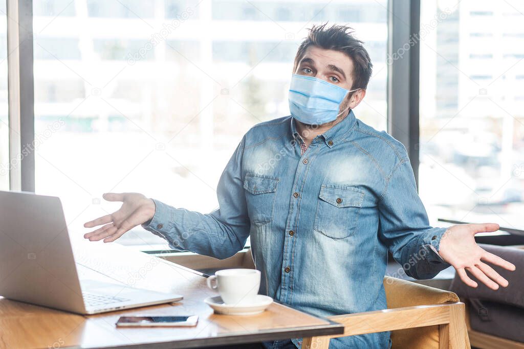Not sure! Portrait of confused young man with medical mask in blue shirt sitting, working on laptop with raised arms and puzzled face, looking at camera. indoor working and health care concept.