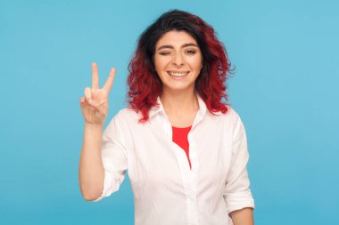 Fantezi kızıl saçlı mutlu hippi kadının portresi kameraya neşe içinde göz kırparak zafer jesti yapıyor, parmaklarıyla barış işareti yapıyor. Mavi arka planda kapalı stüdyo çekimleri