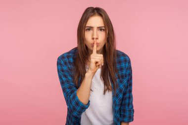 Sus, konuşma! Kareli tişörtlü, dudaklarında parmak tutan, sessiz bir jest yapan, sessiz kalıp sır tutmak isteyen endişeli mutsuz kızın portresi. Pembe arka planda kapalı stüdyo çekimleri