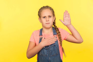 I promise to be honest! Portrait of serious responsible little girl with braid in denim overalls raising palm to take oath, child swearing to tell truth. studio shot isolated on yellow background clipart