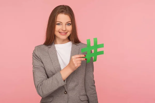 Hash sign. Portrait of elegant blogger, woman in business suit holding paper hashtag symbol, promoting web content and viral internet ideas, blog marketing. studio shot isolated on pink background