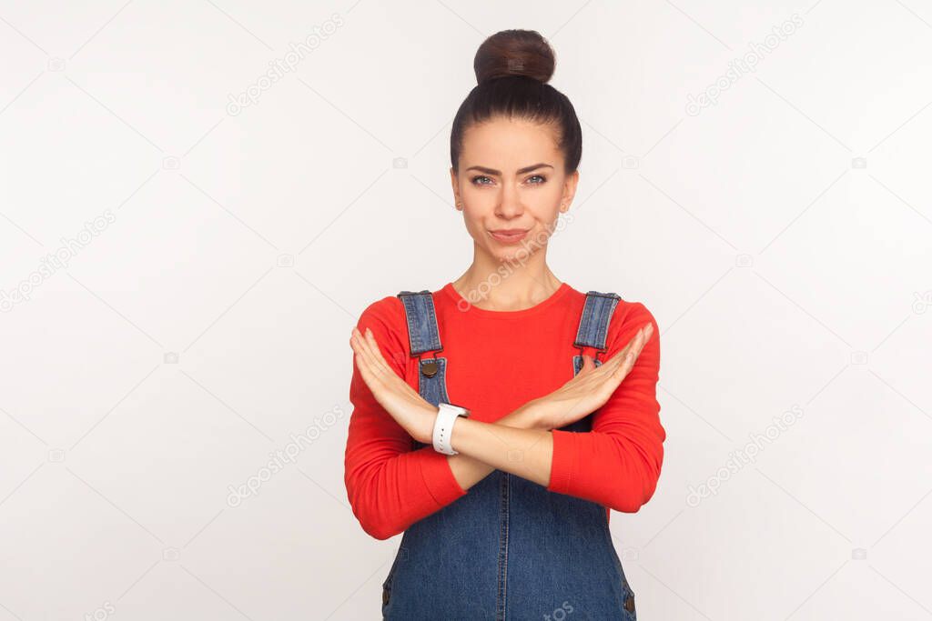 No access! Portrait of strict serious girl with hair bun in denim overalls showing stop sign with crossed hands, rejecting offer, warning expression. indoor studio shot isolated on white background