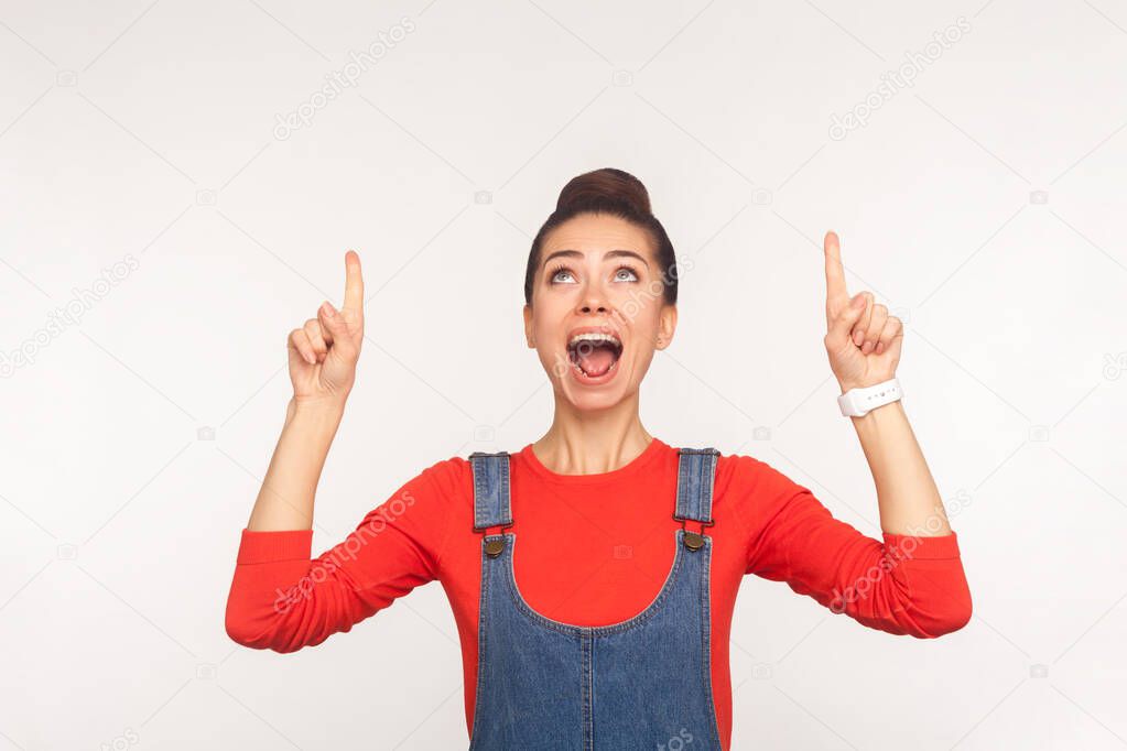 Wow, look at ads! Portrait of surprised shocked girl in denim overalls pointing up at copy space and expressing amazement, showing above blank wall for commercial text. indoor studio shot, isolated