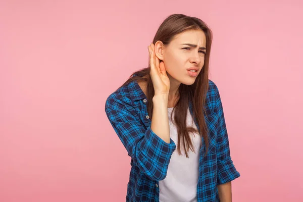What? Repeat I don\'t hear! Portrait of girl in checkered shirt keeping hand near ear, listening to secret rumor with concentrated attentive expression. indoor studio shot isolated on pink background
