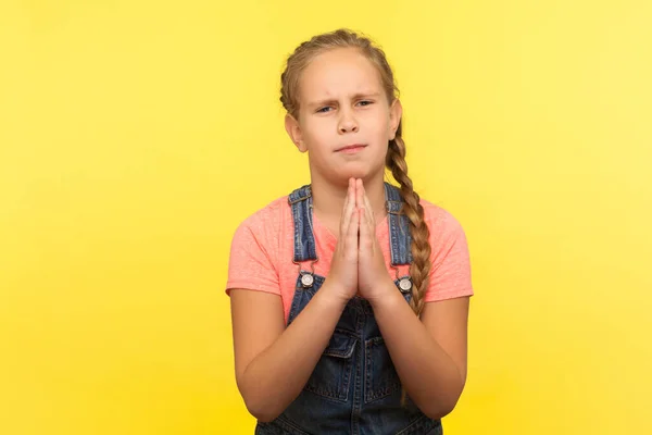 許可を求めて 祈りのジェスチャーで手を握り 許しを請うデニム全体に小さな女の子を埋め込むの肖像画 悪い行動のために謝罪 黄色の背景に隔離されたスタジオショット — ストック写真