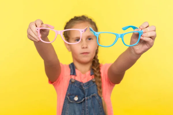 视力和眼睛健康 照片上的小女孩拿着粉色和蓝色的眼镜 表示怀疑 孩子们在光学商店里选择眼镜 在黄色背景下隔离的室内拍摄 — 图库照片