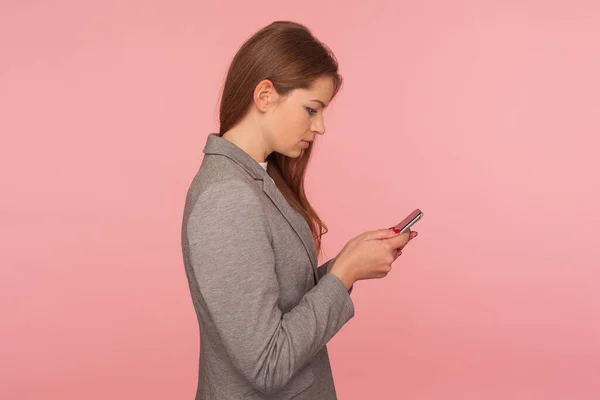 モバイルビジネスアプリ 深刻なのサイドビューは オンラインサービスを使用して スマートフォン上でニュースをチェックし スーツの読書メッセージでエレガントな女性の上司に焦点を当てた ピンクの背景に隔離された屋内スタジオで — ストック写真