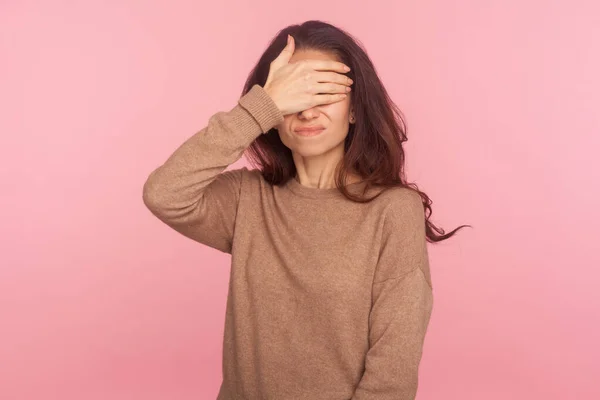 Don\'t want to look! Portrait of young woman hiding her displeased face grimace from something shameful and scary, covering eyes with hand refusing to watch. studio shot isolated on pink background