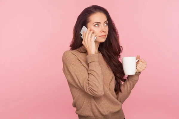 茶色い髪をした若い女性がコーヒーを飲みながらスマホで話している姿を 携帯電話を使って会話を交わしている様子が描かれている ピンクの背景に隔離された屋内スタジオで — ストック写真