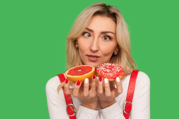困惑的成年女性的衣服 眼睛交叉地看着柚子和甜甜圈 表示怀疑 选择糖果而不是健康的水果 在绿色背景下隔离的室内拍摄 — 图库照片