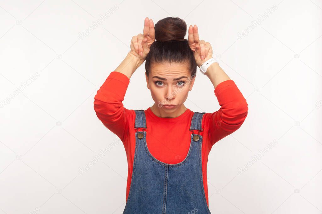 I'm a cow! Portrait of angry girl with hair bun in denim overalls standing with bull horns, antler gesture on her head and threatening to attack, conflict and defence concept. studio shot isolated
