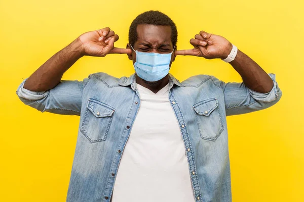 Don't want to hear. Portrait of frustrated irritated man with medical mask covering ears with annoyed expression, ignoring conflict or pressure. indoor studio shot isolated on yellow background