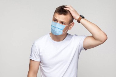 Bir düşüneyim. Cerrahi maske takmış, başını kaşıyarak ne yapacağını düşünen beyaz gömlekli düşünceli genç bir adamın portresi. Gri arka planda izole edilmiş kapalı stüdyo görüntüleri..