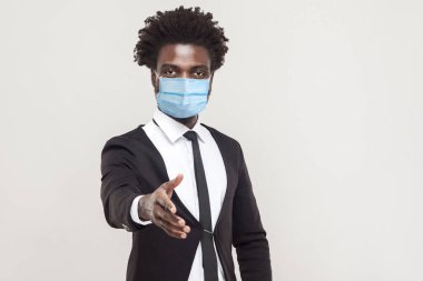 Hoş geldiniz. Cerrahi maske takmış siyah takım elbiseli yakışıklı genç bir adamın portresi. Yardım, destek ya da selamlaşma için ayakta duruyor. Gri arka planda kapalı stüdyo çekimi