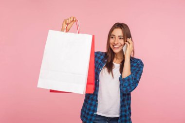 Kareli tişörtlü mutlu kızın portresi. Elinde alışveriş poşetleri, cep telefonuyla konuşmak, satın almak, cep telefonuyla sipariş vermek. Pembe arka planda kapalı stüdyo çekimleri