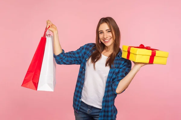 Holiday shopping and presents. Portrait of excited happy beautiful girl in checkered shirt holding packages and gift box, smiling joyfully at camera. indoor studio shot isolated on pink background