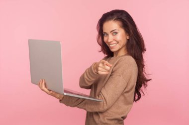 Eline laptop alıp, kamerayı işaret ederek gülümseyen, serbest meslek reklamında seni seçen iyimser genç kadın portresi. Pembe arka planda kapalı stüdyo çekimleri