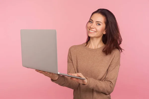 コンピュータ上で活動するフリーランスの肖像画 ラップトップを保持し カメラに笑みを浮かべてカジュアルなセーターの若い女性は コンピュータを使用してオンラインで仕事や勉強 ピンクの背景に隔離されたスタジオショット — ストック写真