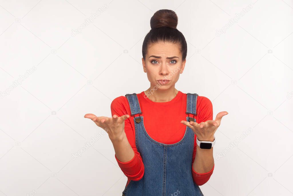 What do you want? Portrait of embarrassed girl with hair bun in denim overalls raising hands with indignant face, asking why how, misunderstanding in conflict. indoor studio shot, white background