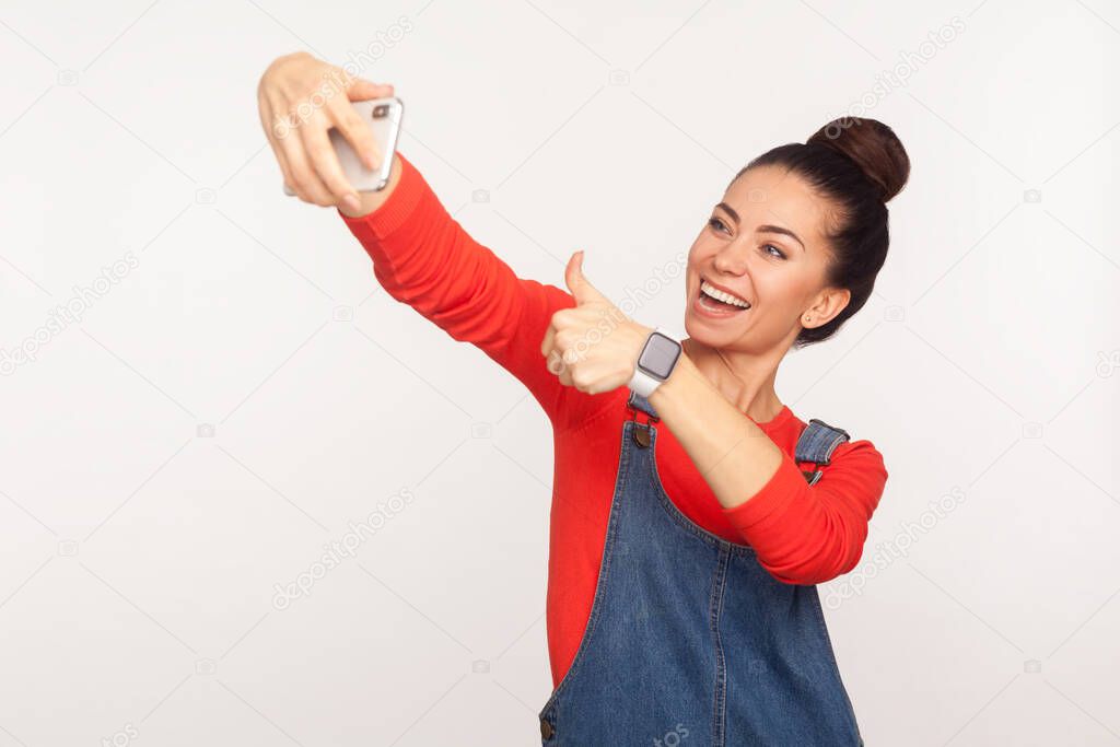 Portrait of cheerful stylish girl with hair bun in denim overalls showing thumbs up, like gesture and making video call on mobile phone, using gadget for online communication. studio shot, indoor