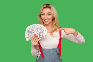 Çok param var! Zengin yetişkin kadın portresi şık tulumlar içinde dolar banknotlarını gösteriyor ve gülümsüyor, nakit depozito ve finansal tasarruflardan memnun. Yeşil arka planda izole edilmiş kapalı stüdyo çekimi