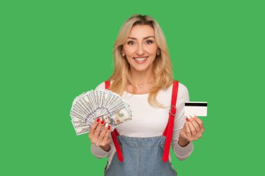 Ticaret, nakit ödeme, elektronik paraya karşı. Dolarlar ve banka kartı tutan, kameraya gülümseyen şık tulumlu mutlu yetişkin kadın portresi. Stüdyo çekimi yeşil arkaplanda izole edildi