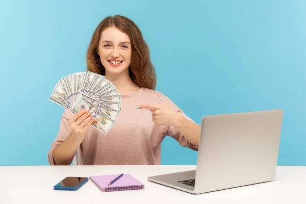 Online betting, earnings on internet. Positive woman pointing at money, freelancer showing us dollar bills, sitting at workplace with laptop home office. indoor studio shot isolated on blue background