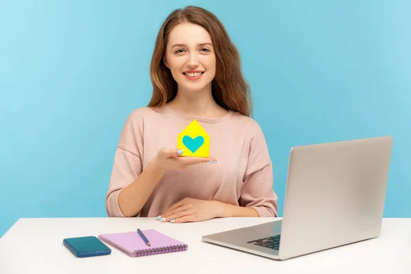 Insurance agency. Cheerful pretty woman holding paper house with heart inside and smiling, real estate agent sitting at desk with laptop, home office. indoor studio shot isolated on blue background