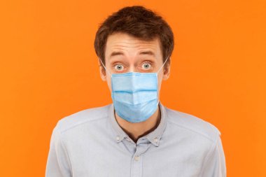 Closeup portrait of shocked young worker man with surgical medical mask standing and looking at camera with big eyes. indoor studio shot isolated on orange background. clipart