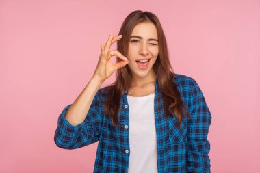 I'm ok! Portrait of cheerful optimistic girl in shirt winking at camera and gesturing okay sign, completely agree with suggestion, confident in success. indoor studio shot isolated on pink background clipart