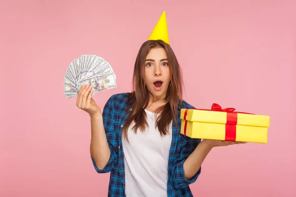 Portrait of happy surprised girl with funny party cone on head holding present box and money, celebrating store anniversary, showing holiday gifts and cashback. studio shot isolated on pink background