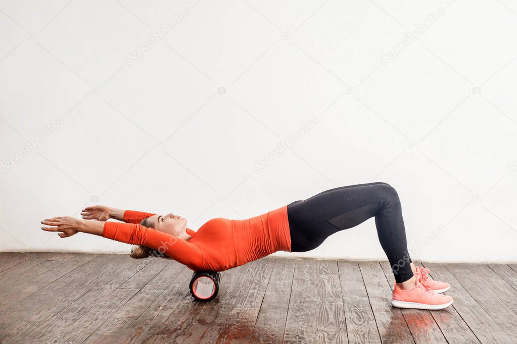 Slim woman in sport tight pants doing exercise with foam roller massager on floor, relaxing and stretching spine muscles, training her back. Health care and workout at home. indoor studio shot