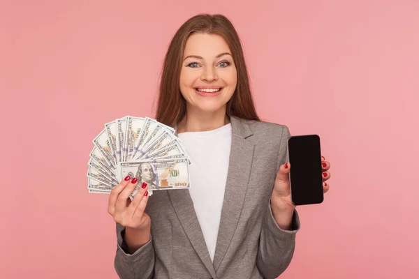 Apply online, loan application. Portrait of happy smiling businesswoman in suit jacket holding dollar banknotes and cellphone, satisfied with mobile banking. studio shot isolated on pink background