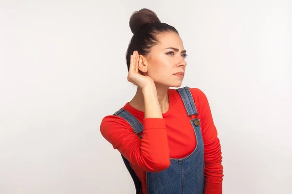 What? I don\'t hear! Portrait of attentive girl with hair bun in denim overalls holding hand near ear listening to gossip, silent talk, confidential information. indoor studio shot, white background