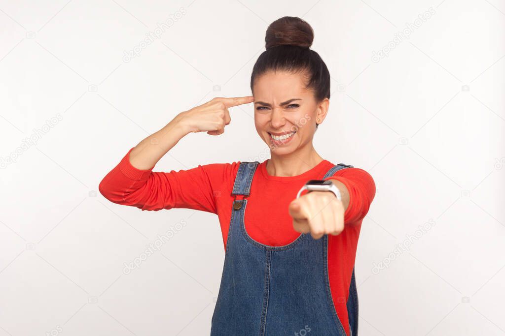 You are dumb, out of mind! Portrait of girl with hair bun in denim overalls showing stupid gesture and pointing to camera, taunting crazy friend with insane idea. indoor studio shot, white background