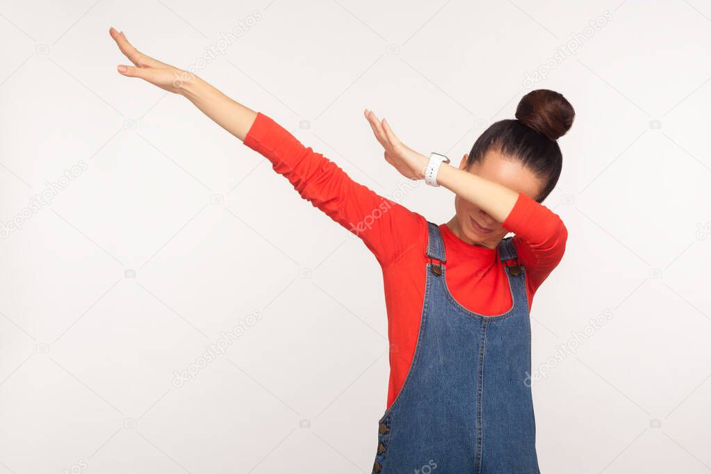 Winning, success gesture. Portrait of stylish girl with hair bun in denim overalls showing dab dance pose, famous internet meme, performing dabbing trends. studio shot isolated on white background