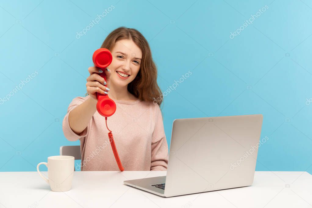 Positive call center operator, woman secretary sitting at workplace and holding old fashioned phone handset, smiling friendly, asking to answer phone. indoor studio shot isolated on blue background