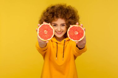 Tüylü kıvırcık saçlı neşeli, hayat dolu kadın kameraya yarım dilim greyfurt, detoks ve sağlıklı çiğ vitamin besin konsepti gösteriyor. Sarı arka planda izole edilmiş kapalı stüdyo çekimi