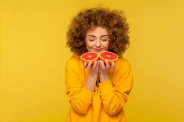 Kıvırcık saçlı, parlak ve mutlu bir kadın yarım dilim greyfurt kokulu lezzetli taze vitamin meyvelerinin ve sağlıklı beslenme düzeninin tadını çıkarıyor. Sarı arka planda izole edilmiş kapalı stüdyo çekimi