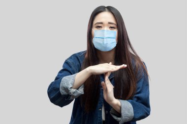 Esmer, Asyalı, cerrahi maske takan, sıradan mavi kot ceketli ve mola tabelasıyla kameraya bakan genç bir kadının portresi. Gri arka planda izole edilmiş kapalı stüdyo görüntüleri..