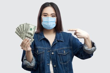 Mavi kot ceketli, mutlu, genç ve güzel bir iş kadını elinde bir sürü dolarla kameraya bakıyor. İçerde, stüdyoda, izole edilmiş, gri arka planda.