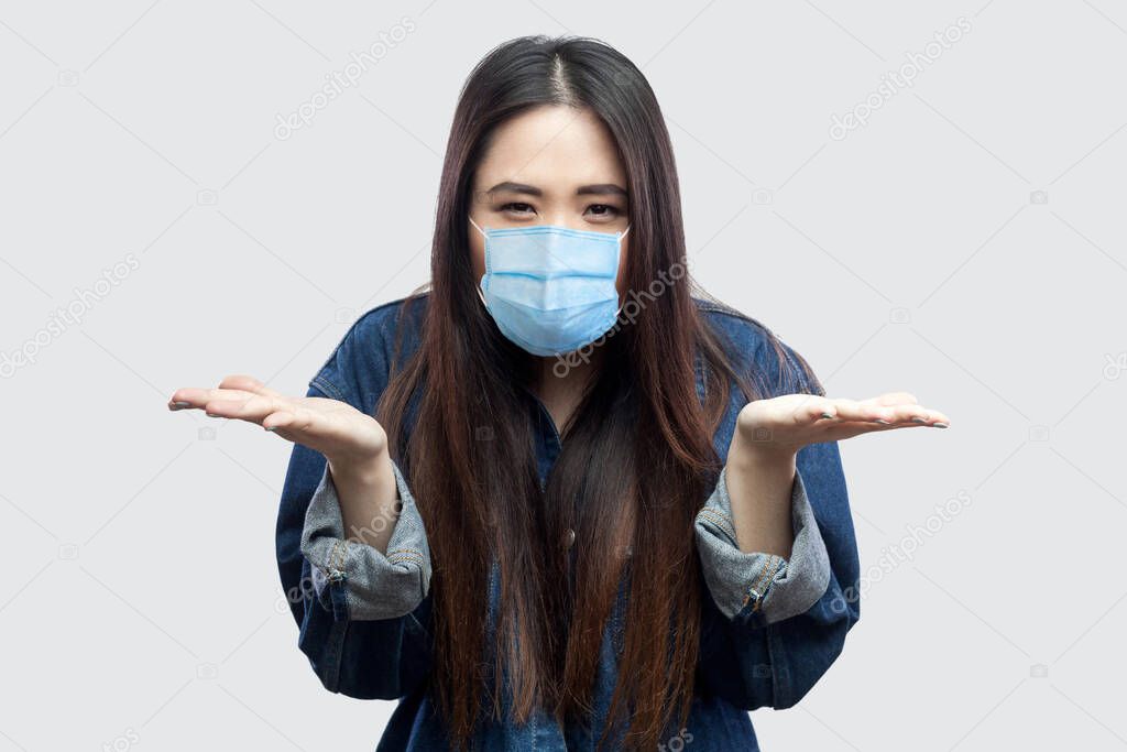 what do you want? Portrait of anger brunette asian young woman with surgical medical mask in casual blue denim jacket standing raised arms and asking. indoor studio shot, isolated on grey background.