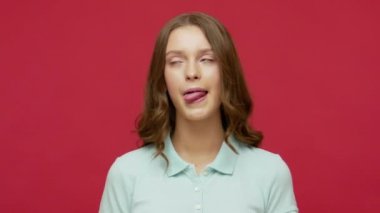 Polo tişörtlü, suratını buruşturup sırıtan itaatsiz edepsiz ifadeyle dilini gösteren, maymun taklidi yapan neşeli, kaygısız genç kadın. Kırmızı arkaplanda izole edilmiş kapalı stüdyo çekimi
