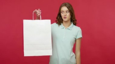 Şaşırtıcı alışverişçi, polo tişörtlü müşteri kadını beyaz fotokopi alanı olan alışveriş torbalarının içine bakıyor, paketleri işaret ediyor ve satın almaktan şok olduğunu ifade ediyor. Stüdyo çekimi izole